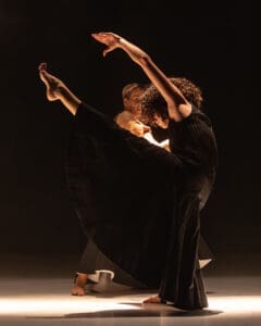 Two dancers create a sculptural duet with extended leg and flowing black costume under theatrical light.