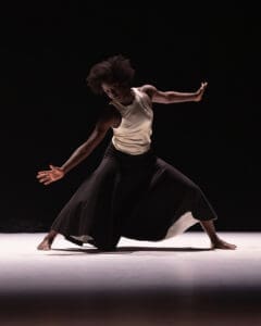 Solo dancer in a grounded contemporary pose with extended limbs under dramatic stage lighting.